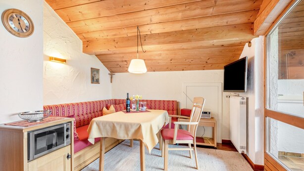 A cozy room with a wooden ceiling, a dining table with chairs, a sofa, a small kitchen area with a stove, and a television. - Apartment 'Ferienwohnung Alpenblick' with Lake View, Private Garden and Wi-Fi (Waging am See-Tettenhausen)