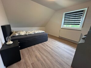 A bedroom with a bed, a nightstand, a lamp, a window with blinds, and a radiator.