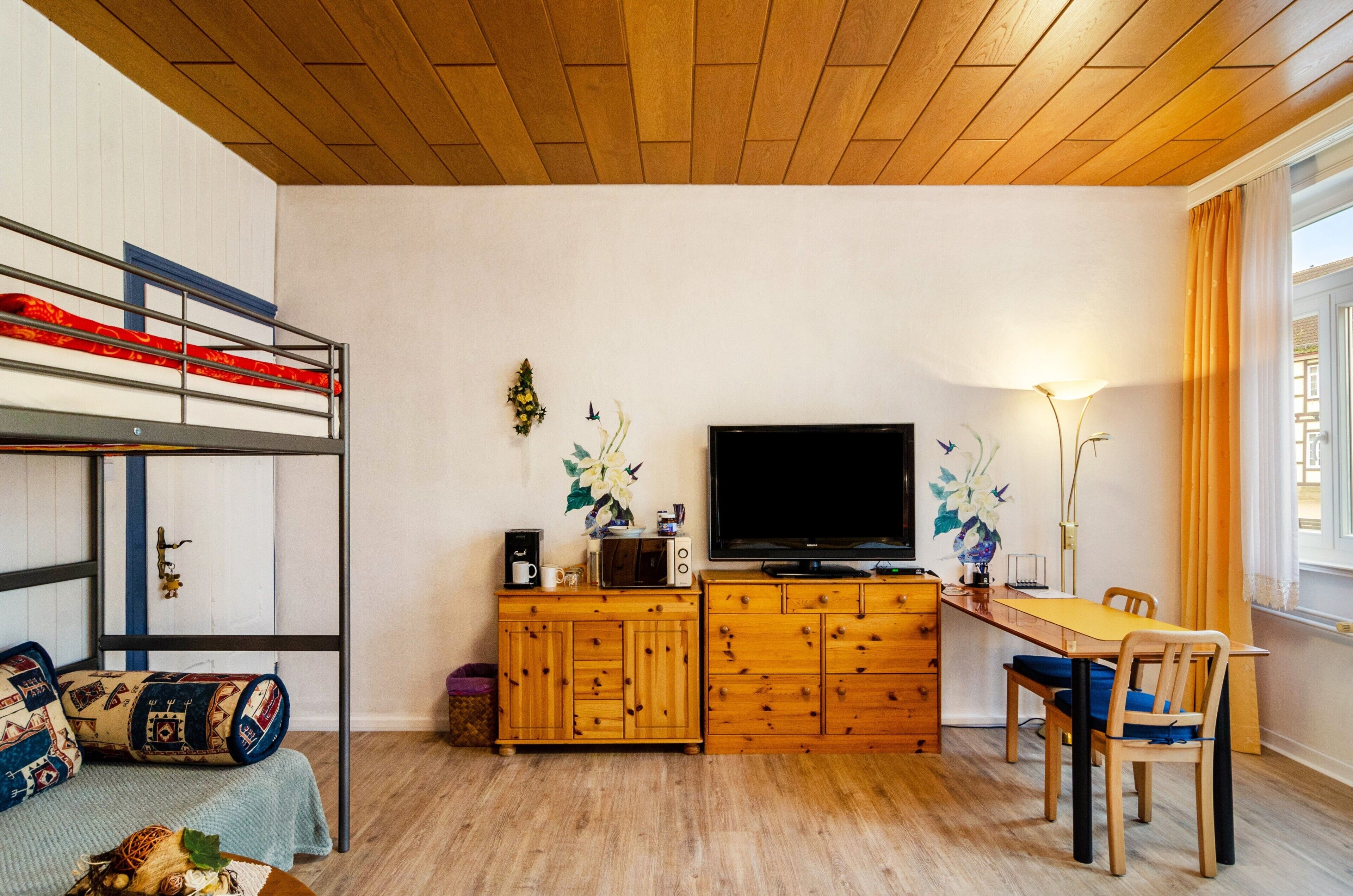 En våningssäng, träskåp, en tv, ett matbord med stolar och ett fönster med gardiner.