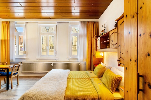 Une pièce comprenant un lit, une armoire en bois, un radiateur et une fenêtre avec des rideaux.