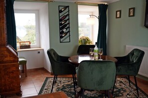 Un coin repas avec une table ronde, des chaises recouvertes d’un tissu vert, un meuble à vin et une fenêtre avec des rideaux.