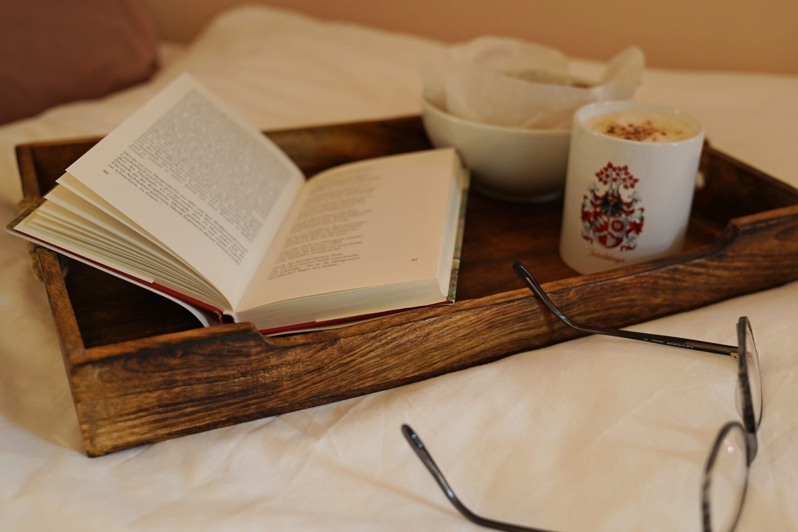 A wooden tray with a book, a mug, and a bowl on a bed.