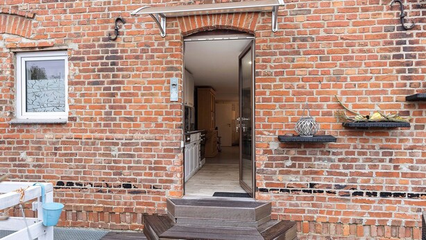 Un bâtiment en briques avec une terrasse en bois, une porte donnant sur un couloir et une petite étagère sur laquelle est posé un objet décoratif.