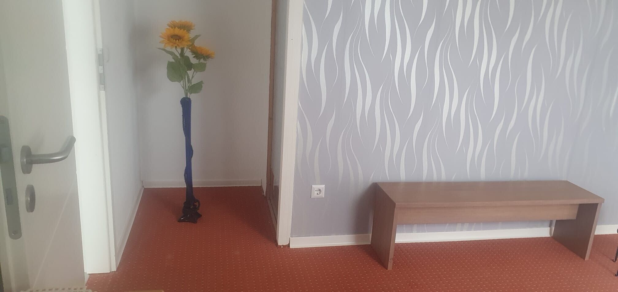 A room with a wooden bench, a vase with flowers, and a patterned wall.