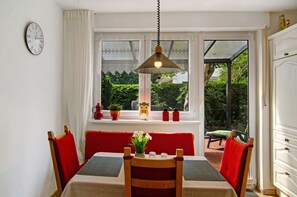 Zona pranzo con tavolo, sedie, lampadario, finestra vista su vegetazione e orologio a parete.