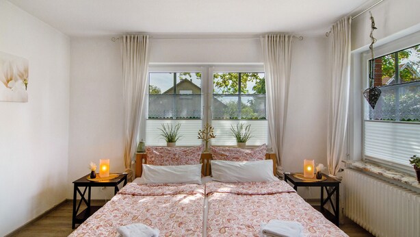 A double bed with a patterned bedspread, two bedside tables with candles, a radiator, and a window with curtains.