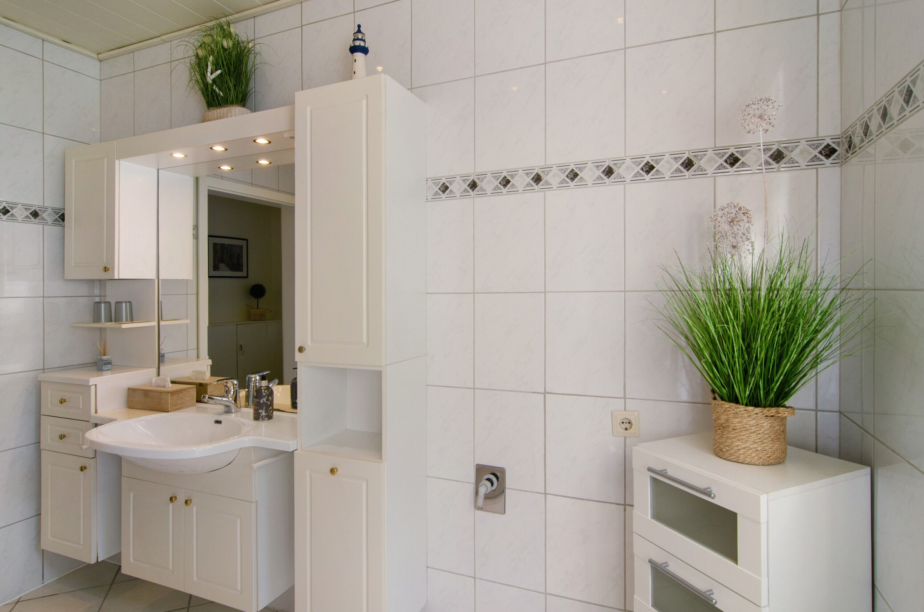Een badkamer met witte tegels, een wastafel, kastjes, een spiegel en een potplant.