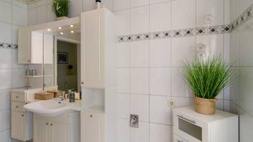 Een badkamer met witte tegels, een wastafel, kastjes, een spiegel en een potplant.