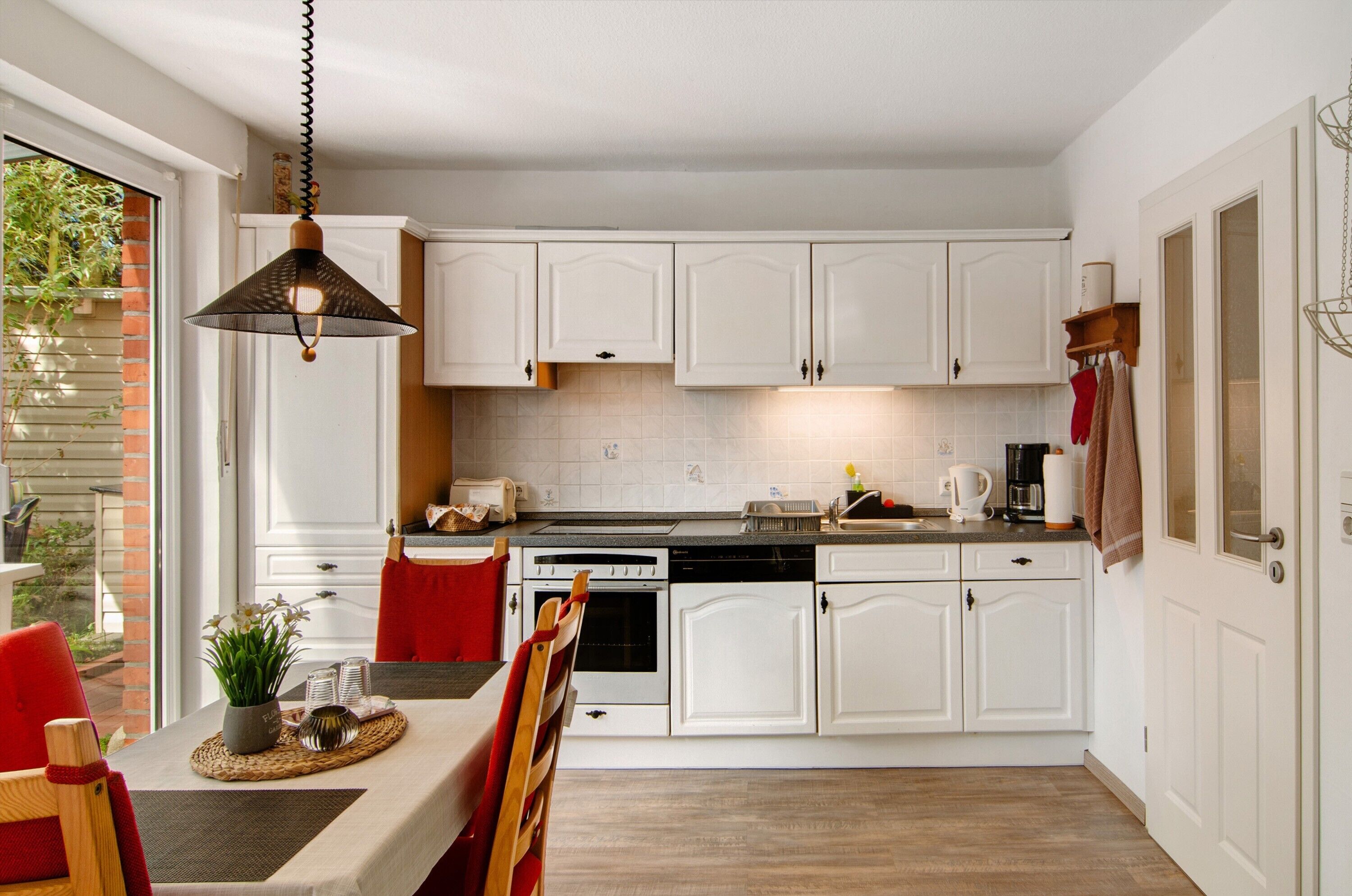 A kitchen with white cabinets, a dining table with chairs, a hanging light fixture, and a view of the outdoors.