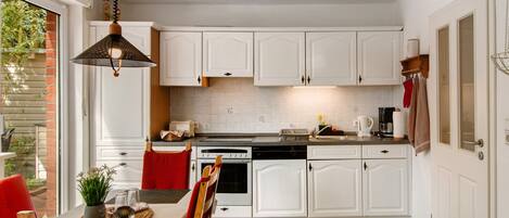 A kitchen with white cabinets, a dining table with chairs, a hanging light fixture, and a view of the outdoors.
