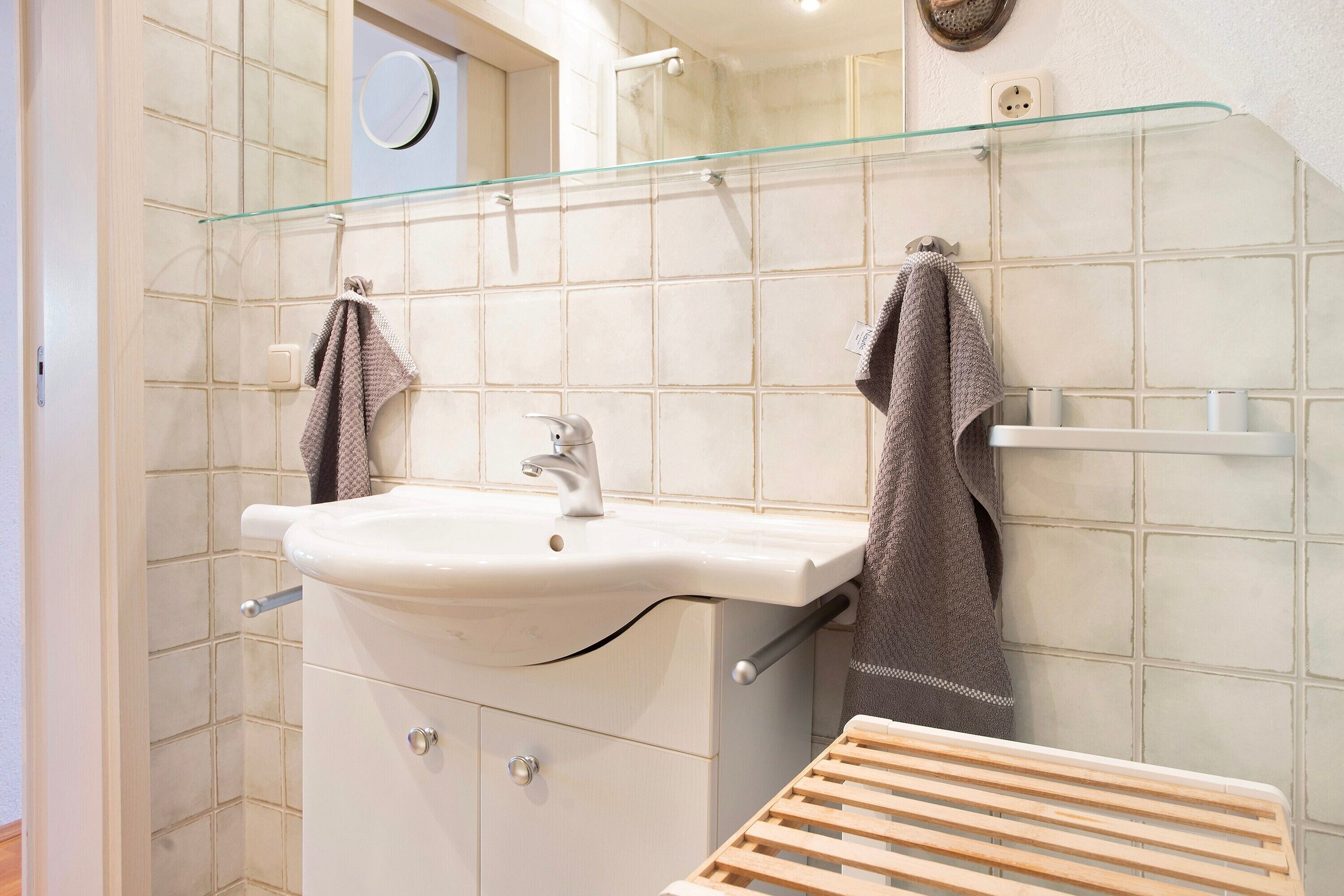 Une salle de bain avec un lavabo blanc, deux serviettes suspendues à des crochets, une étagère en verre et un étendoir à linge en bois.
