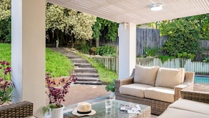 Un espacio de estar al aire libre cubierto con mobiliario de mimbre y una mesa de vidrio, con vistas a un jardín que cuenta con una cerca de madera y un sendero.