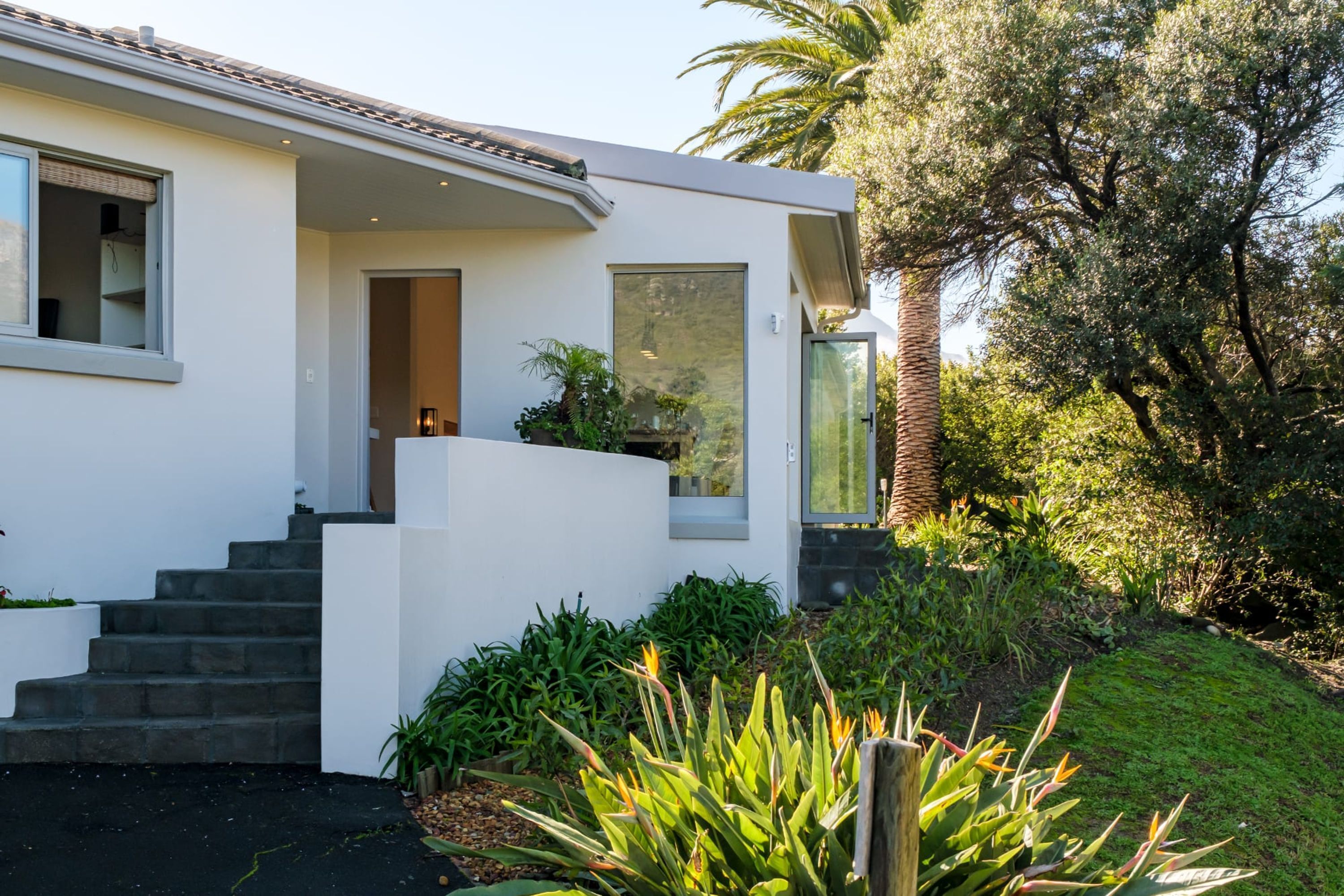 Een modern huis met een witte gevel, een trap die naar een deur leidt, en een tuin met verschillende planten.