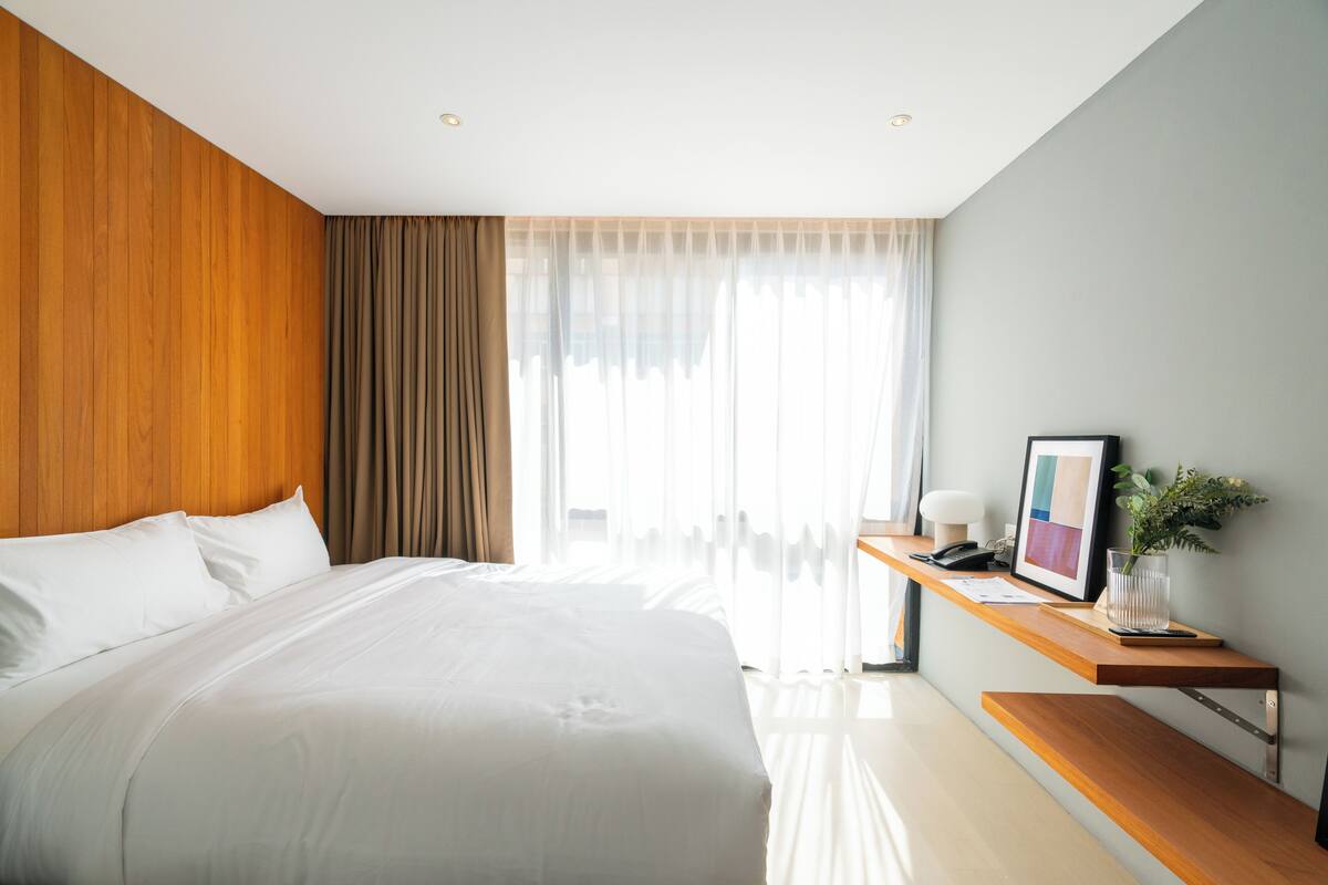 Ein modernes Hotelzimmer mit einem großen Bett, einer Wandpaneele aus Holz, einem Schreibtisch mit Telefon, einem gerahmten Bild und einer Pflanze in einer Vase.