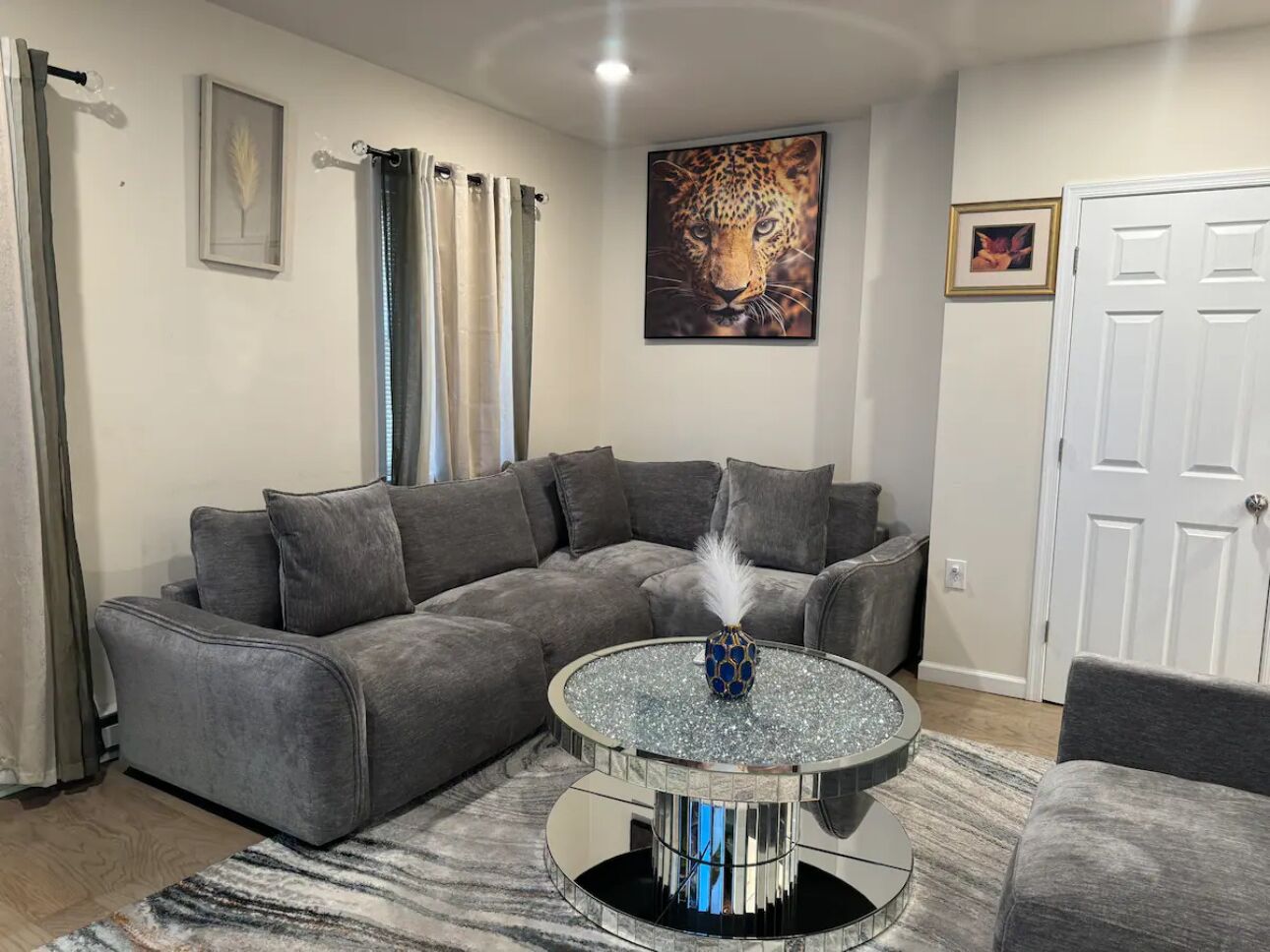 Una sala de estar con un sofá seccional gris, una mesa de centro redonda de vidrio con un jarrón decorativo, y un cuadro enmarcados de un leopardo en la pared.