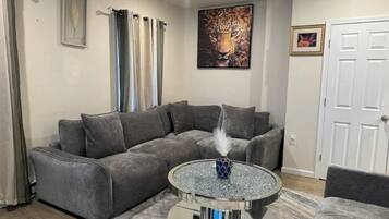 Una sala de estar con un sofá seccional gris, una mesa de centro redonda de vidrio con un jarrón decorativo, y un cuadro enmarcados de un leopardo en la pared.