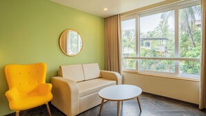 A room with a green wall, a beige sofa, a yellow armchair, a round mirror, a white table, and a large window with a view of greenery. - Bloom Hotel - Dona Paula (Panaji)