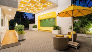 An outdoor area with a yellow wall, potted plants, and seating under an umbrella. - Bloom Hotel - Dona Paula (Panaji)