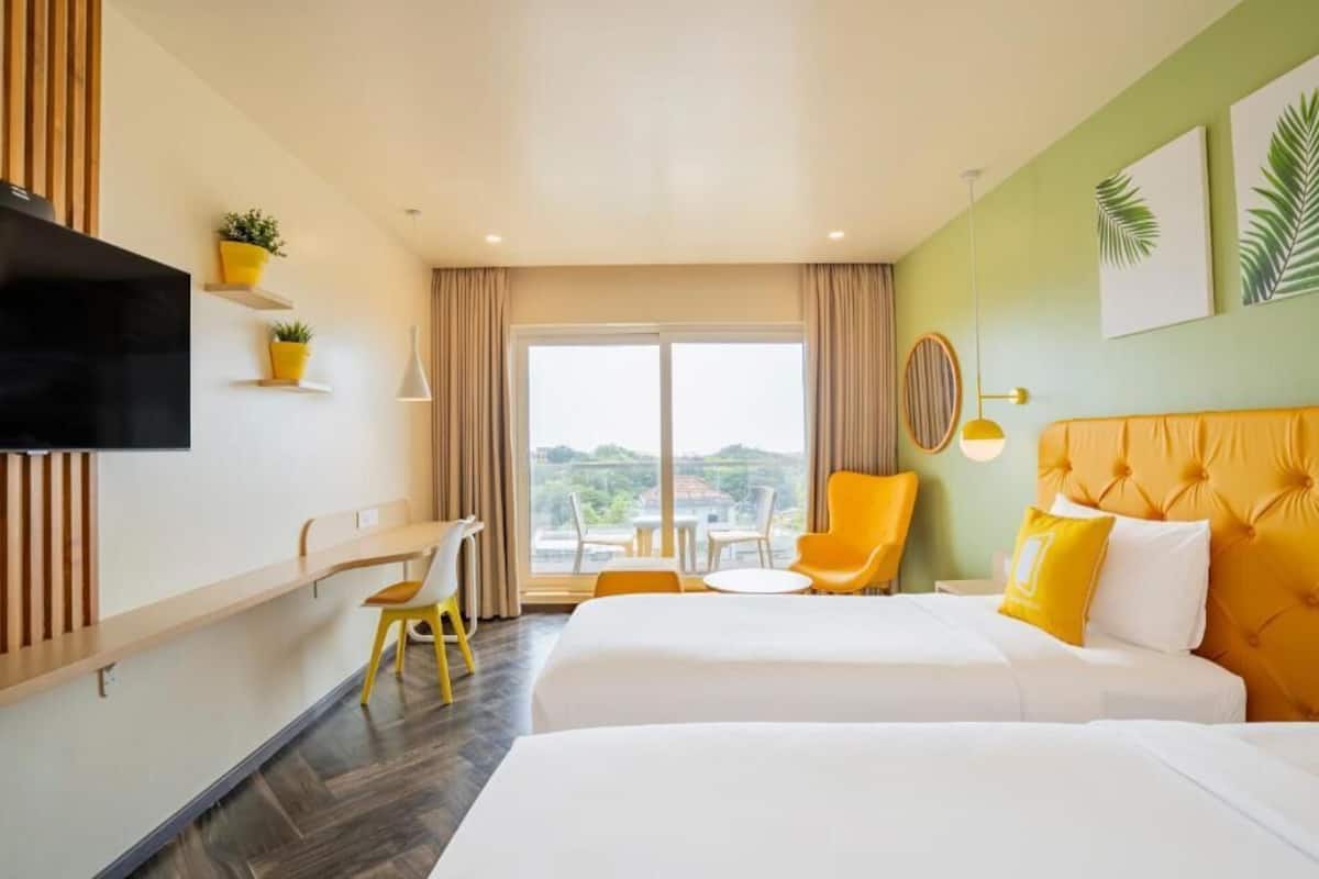 A hotel room with two beds, a desk, a chair, a TV, and a view of the outdoors.