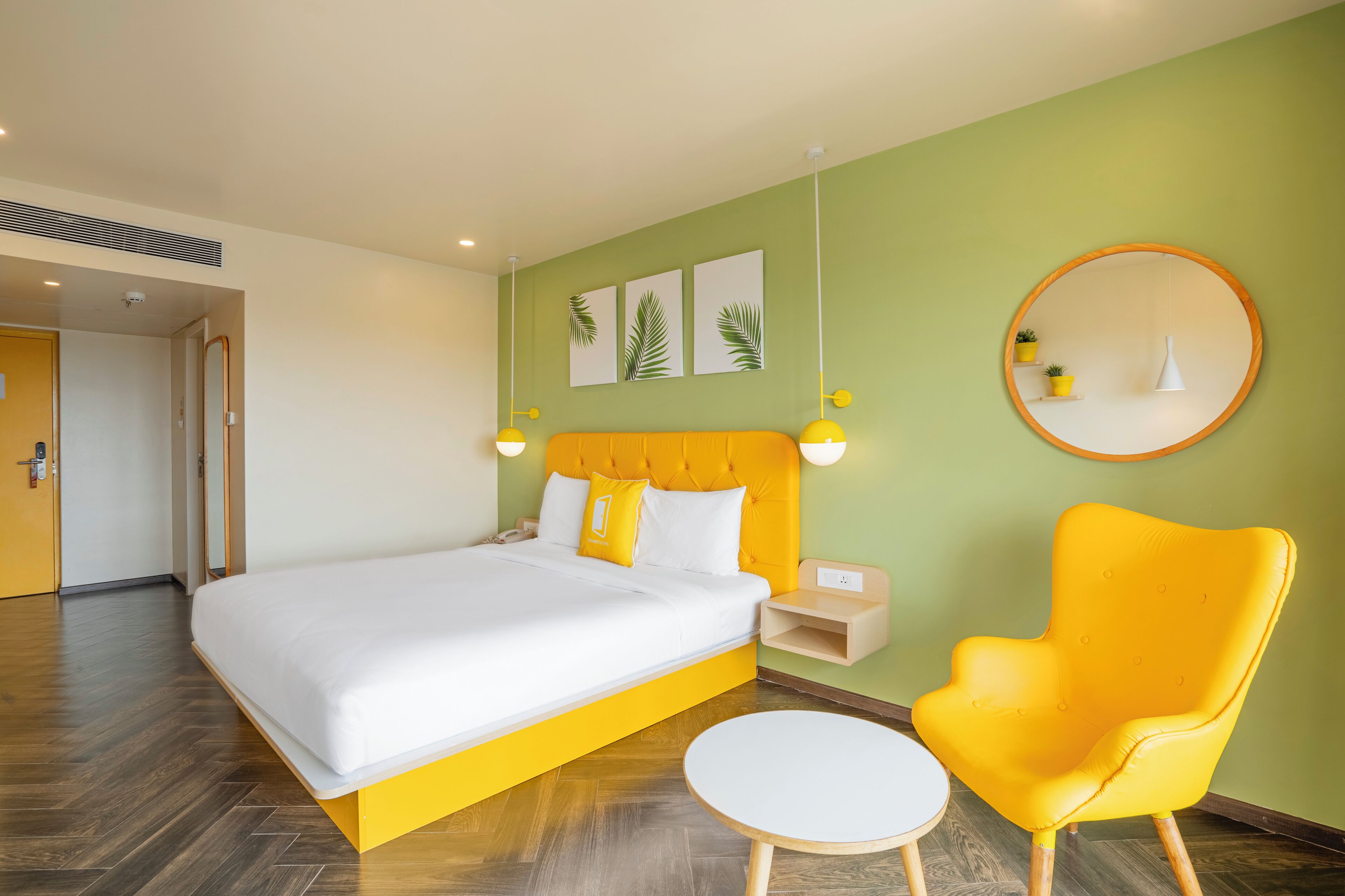 Una habitación de hotel con una cama, un espejo redondo, un sillón amarillo y una pequeña mesa.