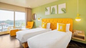 A hotel room with two beds, a large window with a view, a round mirror, and a small table. - Bloom Hotel - Dona Paula (Panaji)
