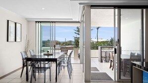 Uma sala de jantar moderna com uma mesa comprida e cadeiras, grandes portas de vidro deslizantes que dão para um pátio externo com vista para a cidade e vegetação.