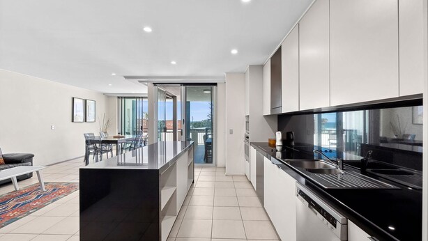 Cozinha moderna com bancada preta e eletrodomésticos de aço inoxidável, área de jantar com mesa de vidro e vista da cidade através de amplas janelas.