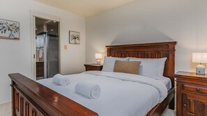 Un lit bien fait, avec du linge de lit blanc et deux serviettes enroulées, une tête de lit en bois, des tables de chevet avec des lampes et une porte donnant sur une salle de bain.