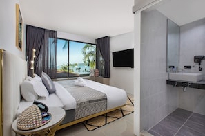 Quarto de hotel moderno com cama, mesinha com produtos de higiene pessoal, banheiro com pia e espelho, e uma janela grande com vista para palmeiras e o mar.