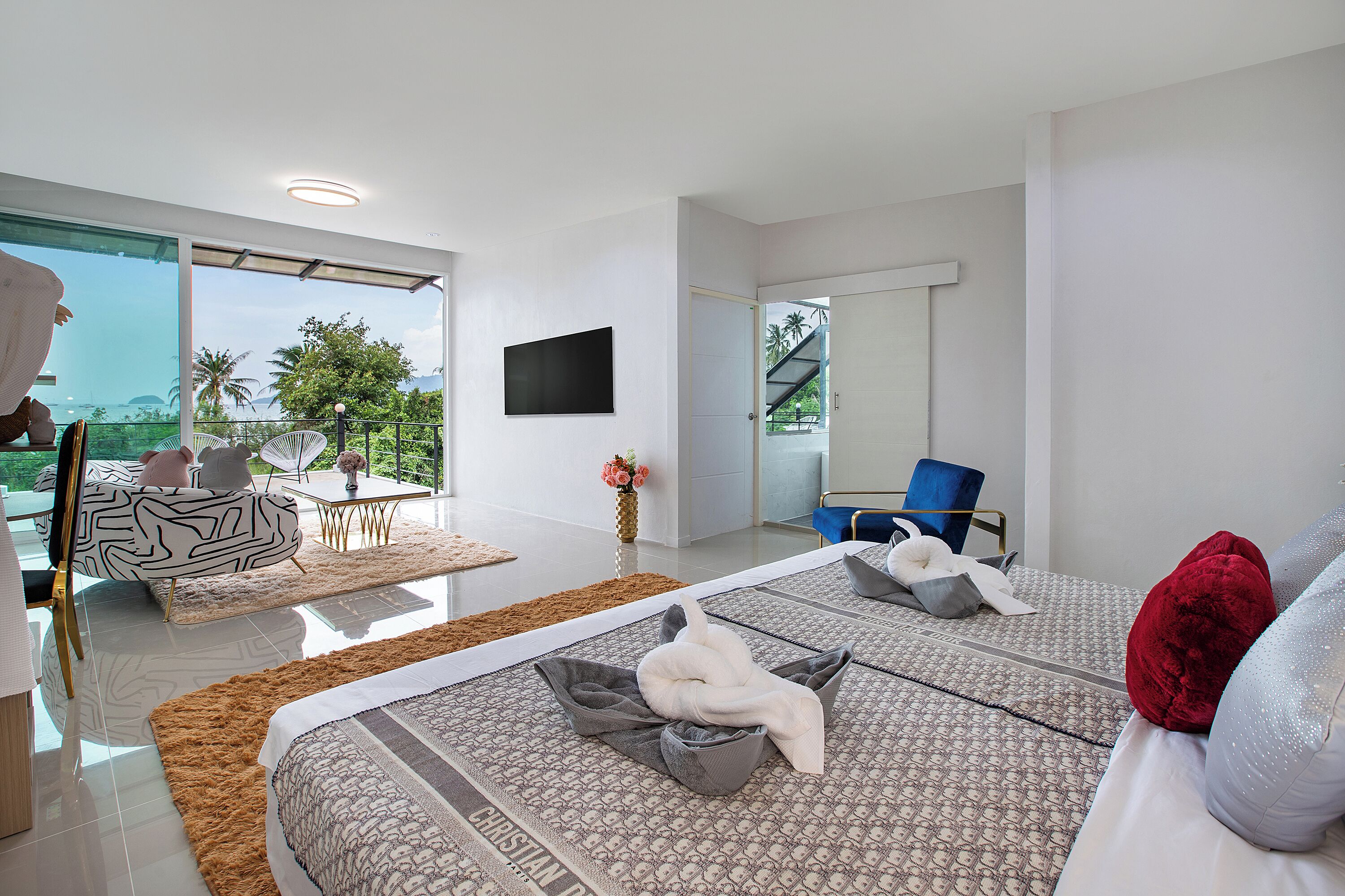 A modern hotel room with a large bed, a dining area with a table and chairs, a TV mounted on the wall, and a balcony with a view of palm trees.