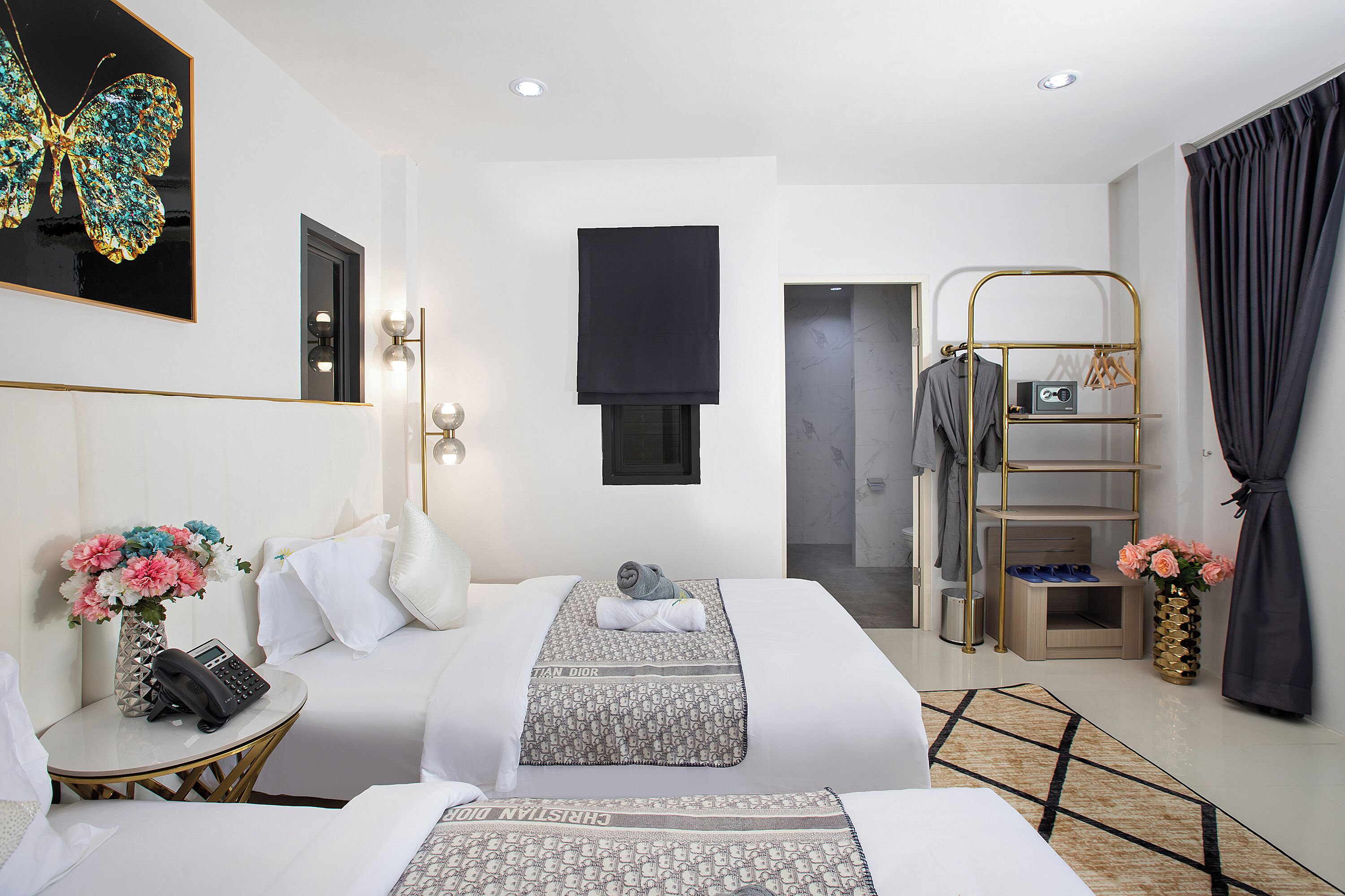 A hotel room with a bed, bedside table, a phone, a vase with flowers, a coat rack, and a mirror.