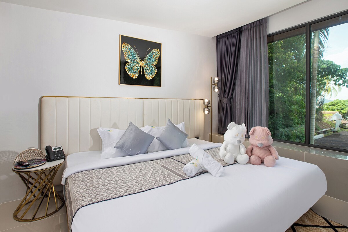 A hotel room with a bed, a bedside table, a telephone, a plush toy, and a window with curtains.