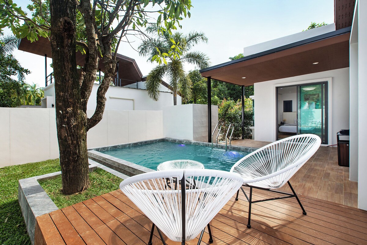 Un espace piscine extérieur moderne, avec deux fauteuils en osier blanc, une terrasse en bois et un arbre.