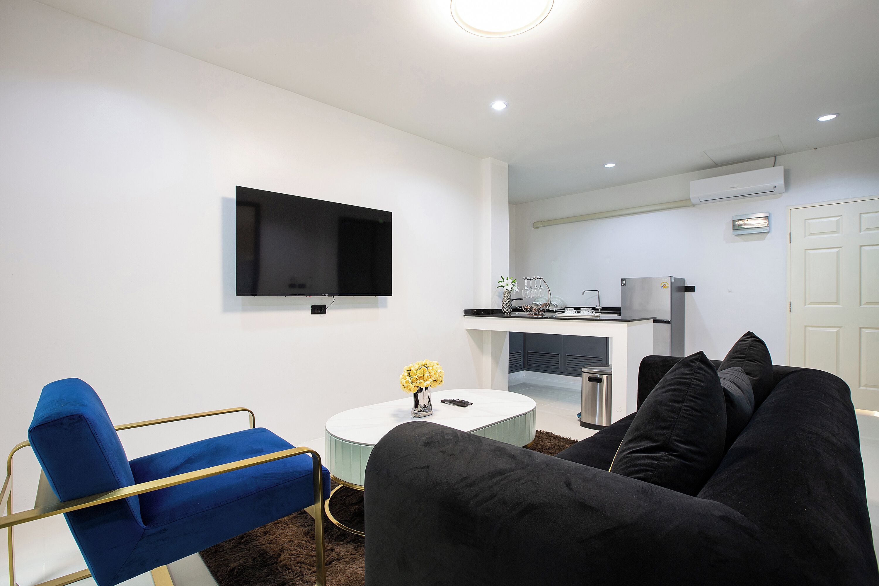 A modern living room with a blue chair, a black sofa, a white coffee table with a vase of yellow flowers, a flat-screen TV mounted on the wall, and a kitchen area in the background.