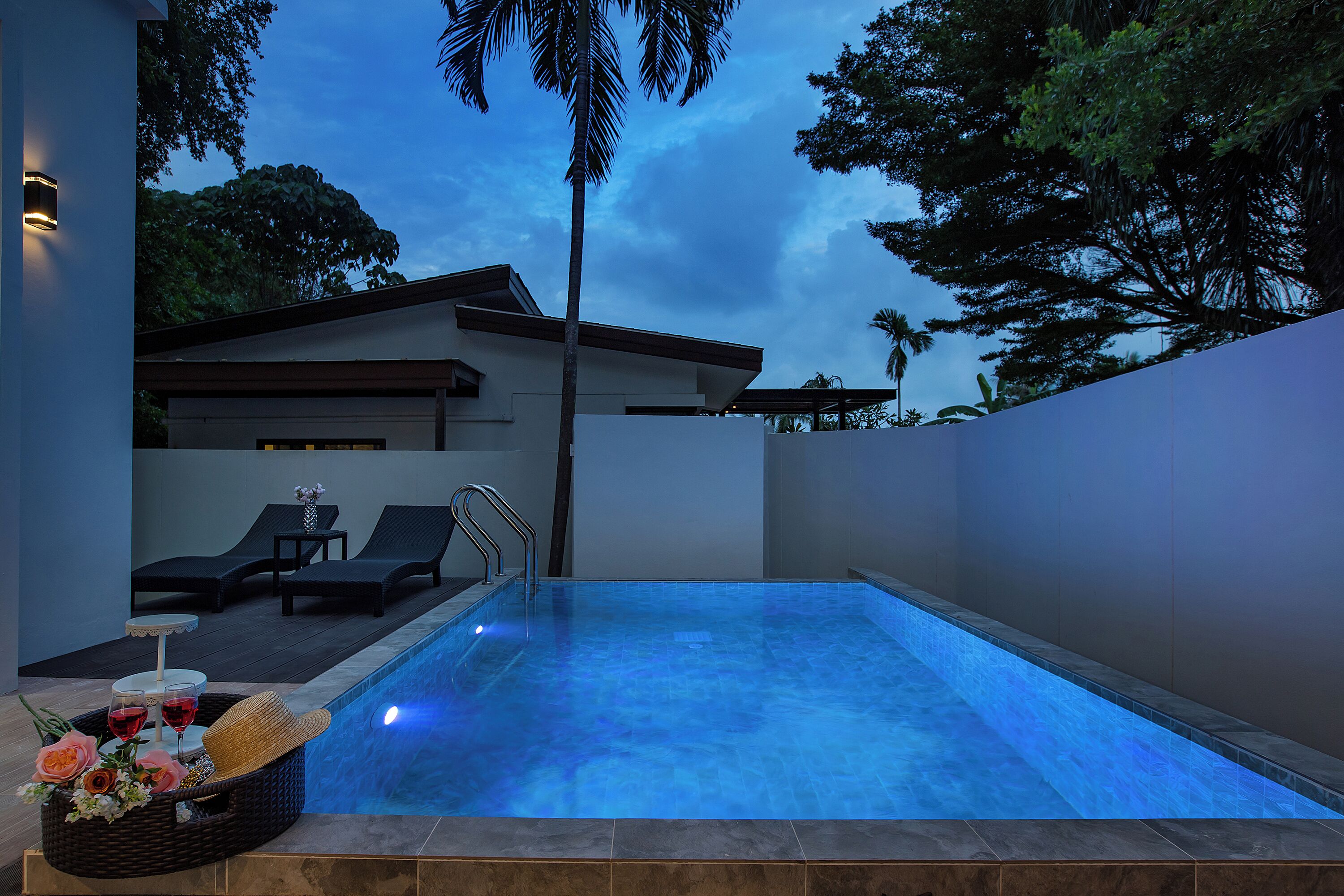 A modern outdoor swimming pool with a blue-lit water feature, lounge chairs, and a small table with drinks and flowers.