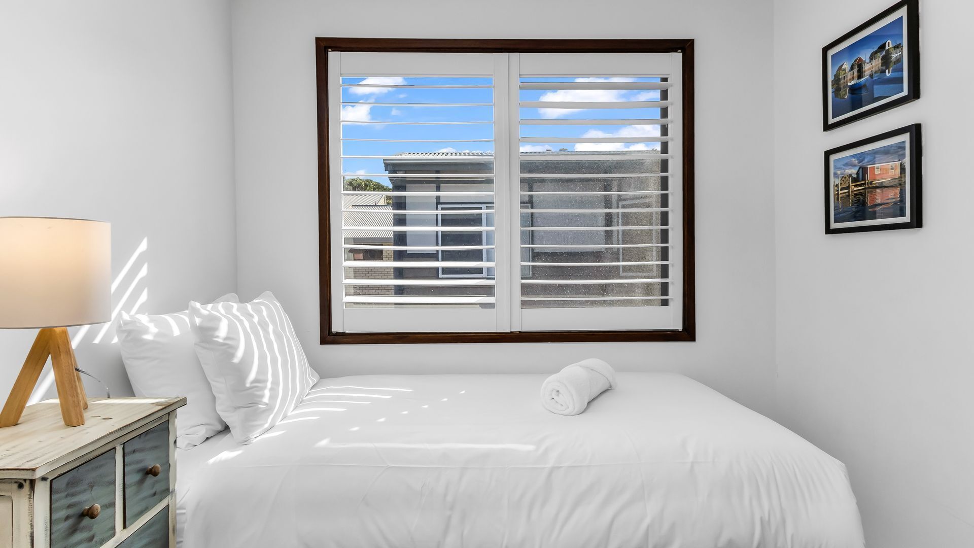 A bedroom with a large window, a bedside table with a lamp, a bed with white bedding, and two framed pictures on the wall.