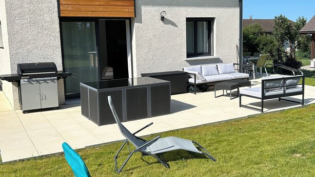 Une maison moderne dotée d’un patio aménagé avec des meubles d’extérieur, un barbecue et une chaise de jardin bleue.