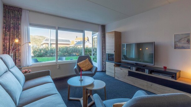 A modern living room with a sofa, a chair, a small round table, a TV, and a window with a view of houses and greenery.