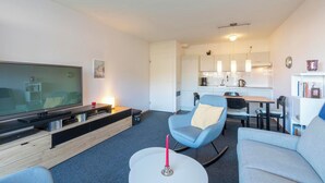 A modern living room with a flat-screen TV, a coffee table, and seating. A kitchen is visible in the background with a dining area.