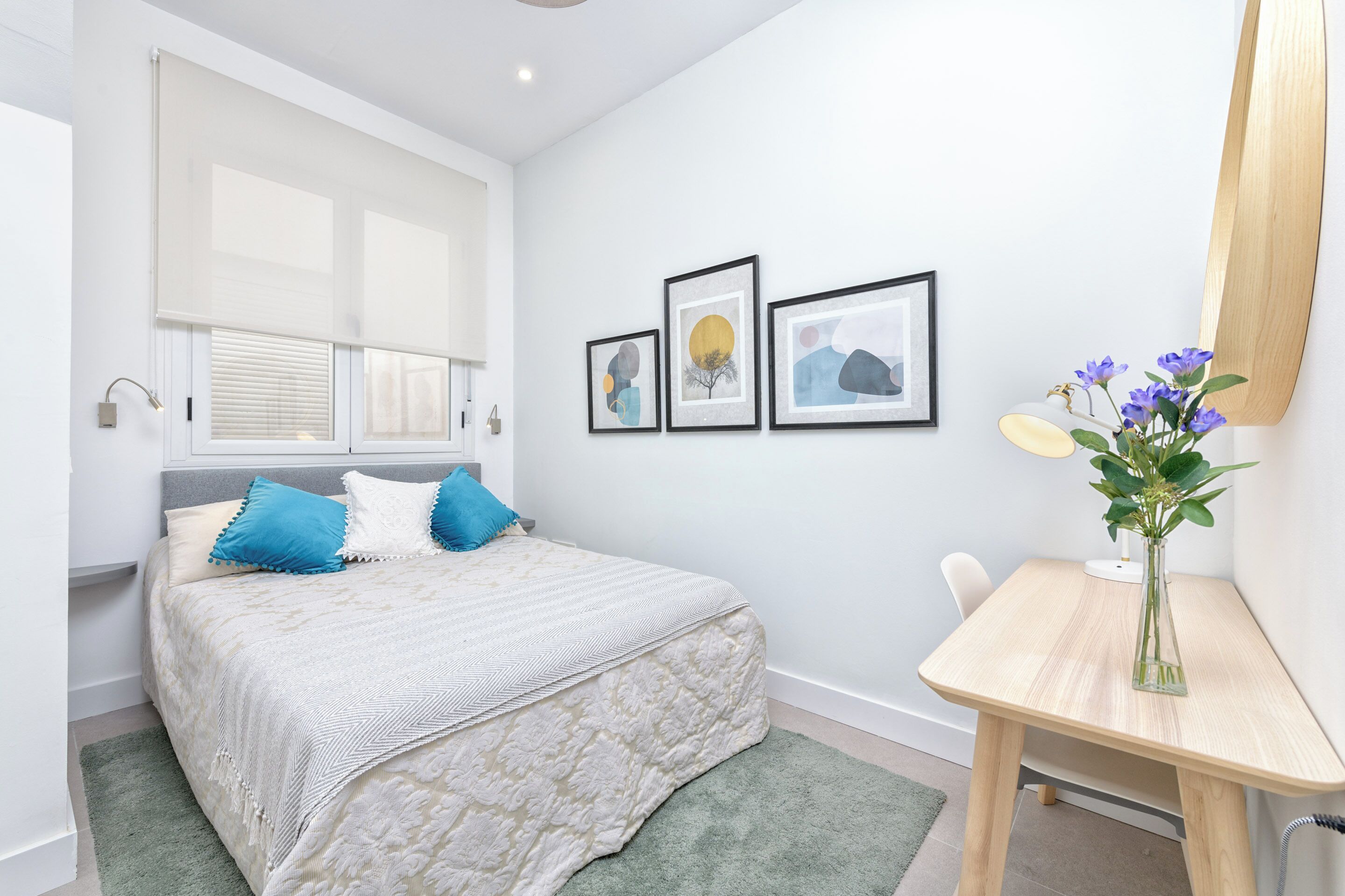 A bedroom with a bed, a desk, and a window with blinds. There are three framed pictures on the wall.