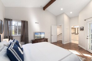 Une chambre avec un grand lit, un téléviseur posé sur une commode, une fenêtre avec des rideaux et une porte donnant sur un coin cuisine.