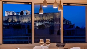 A room with a large window overlooking ancient ruins, two glasses of wine on a table, and modern pendant lighting. - Unique 2BR Penthouse with Stunning Acropolis View (Athens)