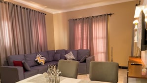 Ein Wohnzimmer mit einer Eckcouch, einem Esstisch mit Unterlagen und einem am Wand montierten Flachbildfernseher.