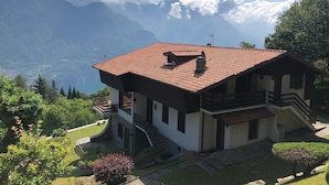 Una casa de dues plantes amb teules vermelles, un balcó i una escala que porta al pis superior. La casa està envoltada de vegetació abundant i muntanyes al fons.