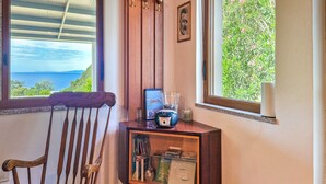 Une pièce d’angle avec une chaise en bois, une petite armoire en bois contenant des livres et une radio, et une fenêtre donnant sur un paysage verdoyant et la mer.