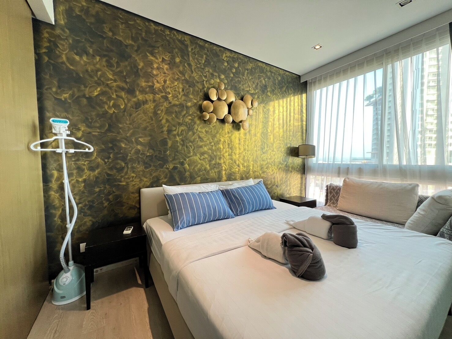 Ein modernes Hotelzimmer mit einem großen Bett, einem Wandheizkörper, einem Nachttisch mit Lampe und Blick auf die Stadt durch große Fenster.