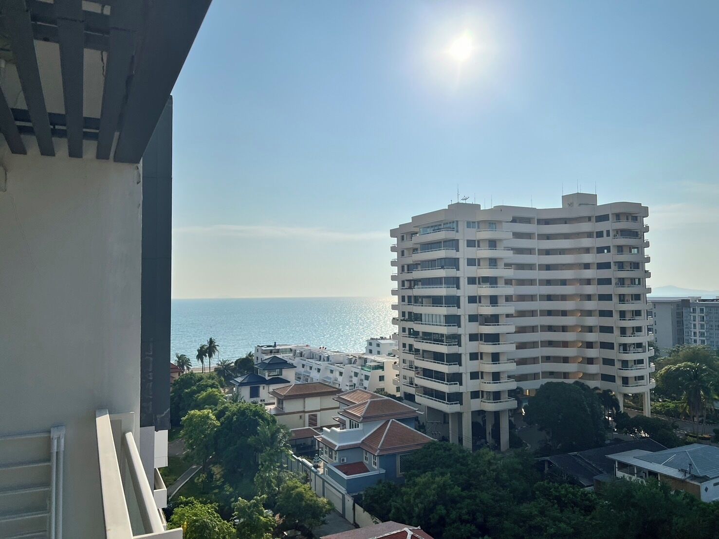 A high-rise building with multiple balconies, surrounded by residential houses and palm trees, overlooking a body of water.