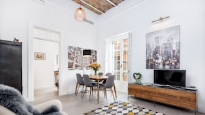 A modern living room with a sofa, a dining area with a table and chairs, a TV on a wooden cabinet, and a cityscape painting on the wall. - Solaga - Casapalma (Málaga)
