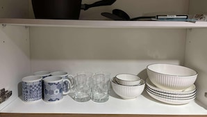 A kitchen shelf with a black pot, a spatula, and a pen. Below, there are two patterned mugs, several clear glasses, and a stack of white bowls with black stripes. - Chic 1-bedroom apartment in brilliant Pasay with WiFi, AC (philippines)