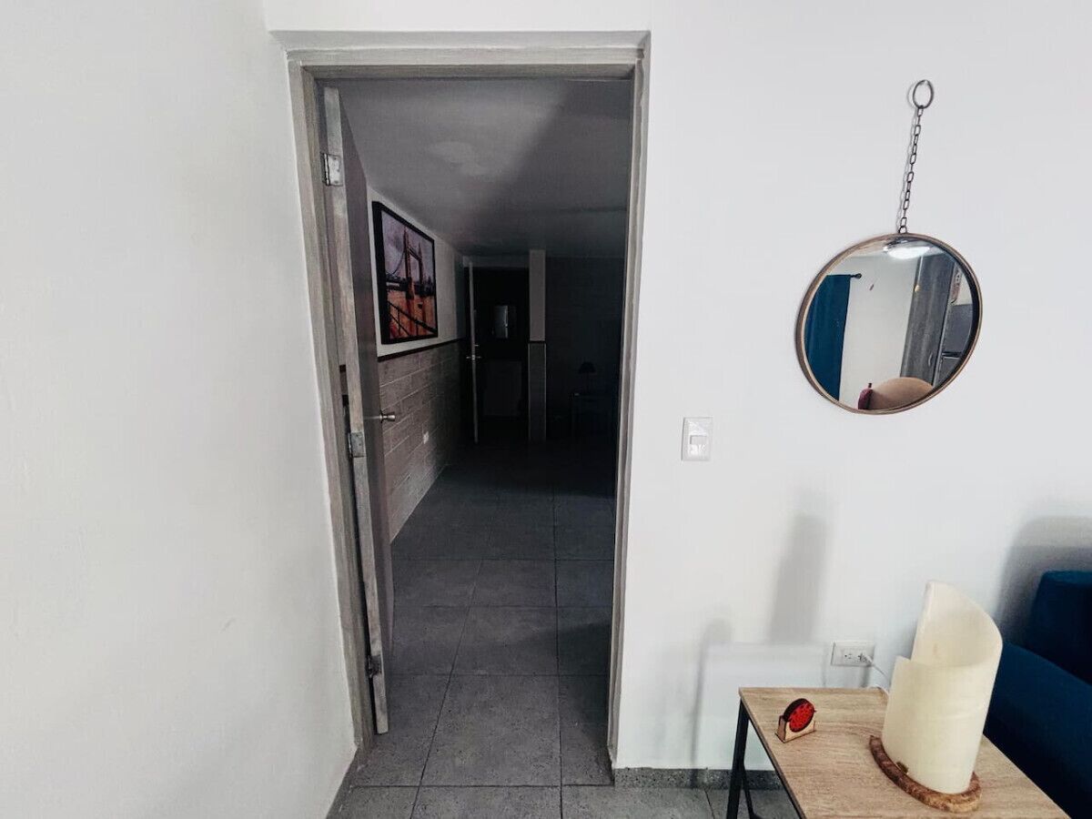 Un pasillo con una puerta que da a una habitación, un espejo en la pared y una pequeña mesa con una lámpara y un objeto rojo.
